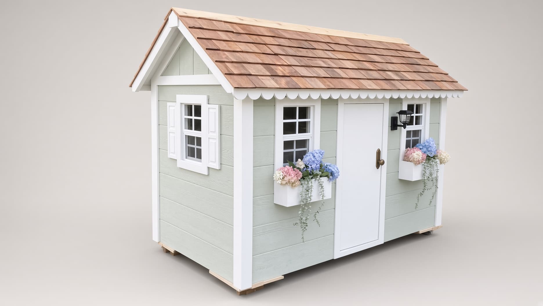 A handcrafted sage-green Little Dream House playhouse with a cedar shingle roof, scalloped white trim, and hydrangea window boxes